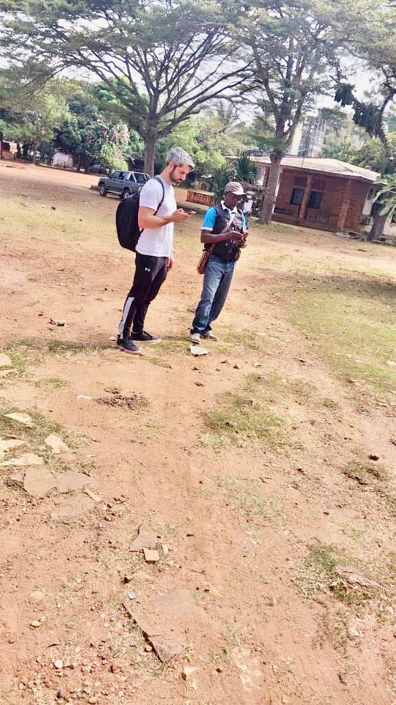 Two men stand in a grassy, open area with trees and buildings in the background, both looking at their phones.