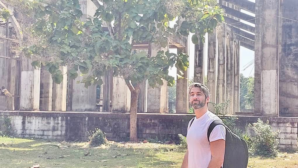 A man with a backpack stands in front of a crumbling structure with large columns, partly obscured by a tree.