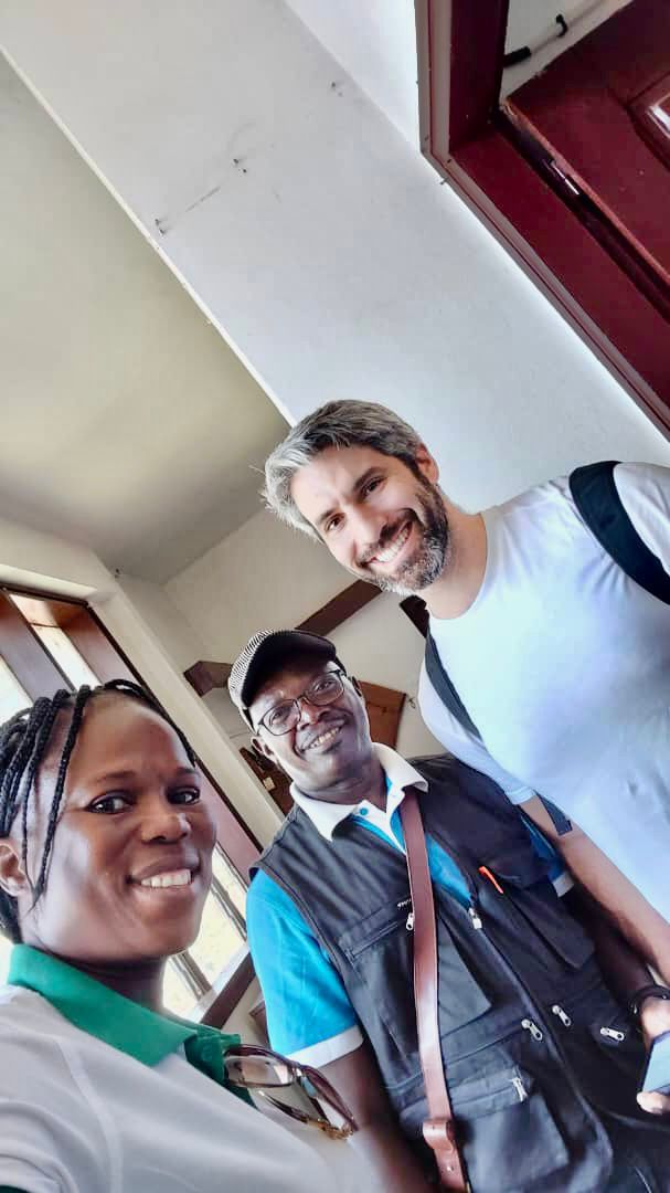A selfie of three smiling people in a hallway, with a woman in a white and green shirt, a man with glasses and a hat, and a man with a beard and a white shirt.