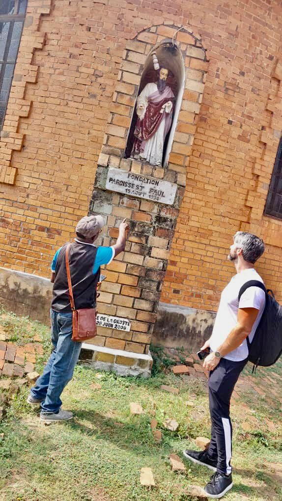 Two men are standing outside a brick building, one pointing at a sign on the wall that reads